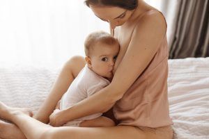 young-tender-mother-in-sleepwear-breastfeeding-hugging-her-newborn-baby-sitting-in-bed-at-morning